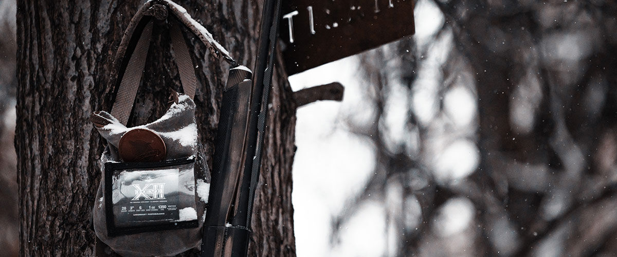 HEVi-XII box hanging from a shotshell bag on a tree next to a shotgun in the snow