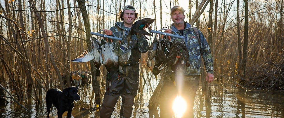 two hunters standing in water holding ducks next to a dog