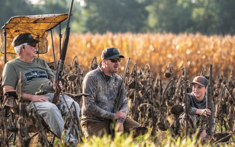 3 generations of hunters kneeling or sitting in front of a field of dried corn